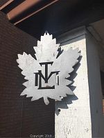 Metal silver maple leaf wall hanger with TML cut-out letters hanging on exterior wood post under roof eave.