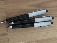 Three black and silver retractable gag gift pens placed on a wooden surface showing pens in a stacked position.