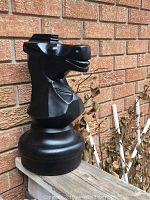 Large black plastic knight chess piece shown outdoors against brick wall from an angle revealing head and upper body details.