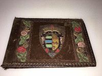 Photo of a brown leather antique photo album cover with embossed rose patterns and a multicolored heraldic crest in the center, edges have stitching and visible wear.