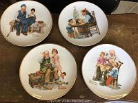 Four Norman Rockwell decorative plates arranged on a flat surface, showing full plate artwork and gold rim detail.