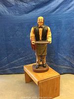 Full front view of vintage wooden butler statue standing on a small wooden table against a blue backdrop.