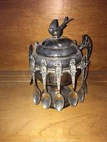 Front side view of the silver plated Victorian spooner sugar bowl with bird finial lid and six spoons hanging around it. One handle missing can be seen on the right side.