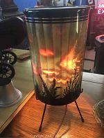 Full view of vintage motion lamp illuminated on tripod stand showing nature silhouette and striped shade pattern, some internal damage visible.