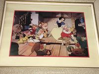 Framed Disney lithograph showing Snow White dancing with the Seven Dwarfs inside the cottage, beige textured frame visible.