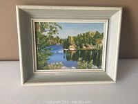 Frontal view of the oil painting showing a riverside landscape with trees and water, in white frame.