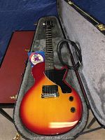 Cherry red guitar body inside grey plush-lined case with cable and strings pack visible