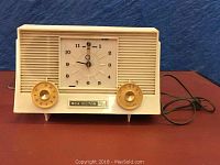 Front view of vintage RCA Victor radio with clock, showing plastic beige casing, speaker grill, clock face and tuning/volume knobs, powered by electric cord.
