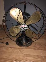 Front view of antique Peerless electric fan with brass blades and protective cage, wire visible on left side.