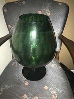 Photo of a large dark green bulbous glass vase sitting on a gray floral upholstered chair, showing front angle and overall size and shape.
