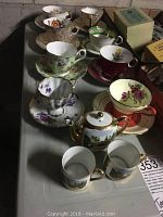 Multiple teacups and saucers of various floral designs and colors arranged on table alongside small child-sized teapot and mugs.