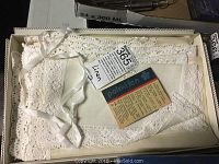Box containing folded white linen tablecloth with lace edging and a tag labelled 'Linen'