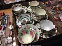Top view of assorted teacups and saucers with floral designs and gold trim in a box, with medals visible nearby
