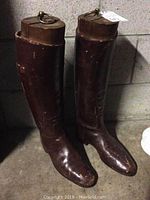 Full view of the pair of brown leather tall boots upright against a grey concrete wall, showing the full length and wooden shoe trees inserted.