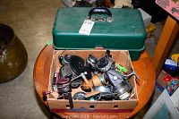 Green plastic fishing tackle box closed with box of fishing reels in cardboard box in front, placed on a small wooden round table