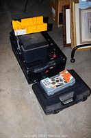 Two stacked black plastic toolboxes with a small yellow organizer and a black plastic case containing hand tools on top, items arranged on a concrete floor near some framed pictures.