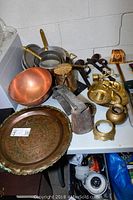 Copper pot, copper platter, brass candle holders, cow bell, decorative sword, wood and brass decor items on a table against a cinder block wall.