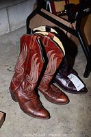 Two pairs of cowboy boots side by side, one lighter brown with wing-shaped design, other darker brown. Showing wear and scratches.