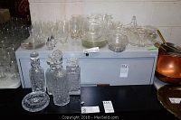 Photo showing various cut glass decanters and an ashtray on a black surface in front of clear glasses and plates.
