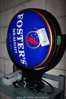 Front view of round Foster's Draught sign mounted on bracket with power cord visible.