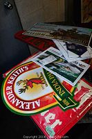 Photo shows various vintage beer and sports signs stacked on a table, including a large Alexander Keith's India Pale Ale metal sign and other beer advertising signs.