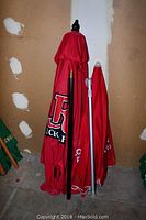 Two red Rickard's patio umbrellas folded up, showing one with a black pole with finial and the other with a silver pole with crank