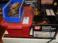 Wide view showing blue bin with loose nails, red organizer bin with screws, box of Grip Rite framing nails, and four staplers on top.