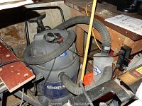 Blue and gray 20 gallon Shop Vac with attached long hose under a workbench.