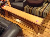 Long cedar and walnut wooden bench with crossed legs on wooden floor surrounded by furniture.