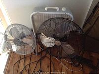 Photo showing all three fans placed on the floor: a white box fan in the back, a black pedestal Utilitech fan in the middle, and a silver Cool Breeze pedestal fan on the left.
