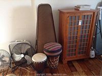 Overall view of cello case, two fans, wooden cabinet and two drums on wood floor