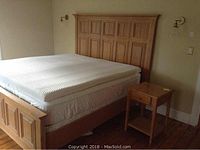 Overall view of light brown king size bed frame with mattress and matching side table next to it in a bedroom with wood flooring.