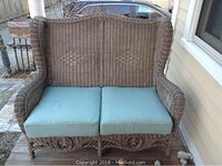 Front view of the tan wicker settee with turquoise cushions on a wooden porch floor.