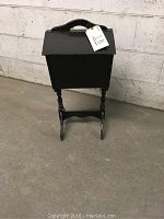 Front view of black antique sewing stand with dual flip lid tops and wooden wheels, placed against a wall.