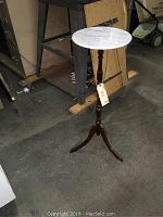 Full view of the plant stand showing marble top and wooden tripod base in an indoor storage setting.