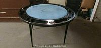 Round Durham Line folding poker table set up with blue felt top and black plastic tray with cup holders and chip slots around the edges, supported by foldable legs.