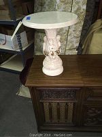 Full view of the plaster cherub base side table with round white marble top, sitting on a wooden cabinet.