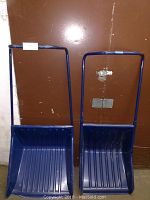 Two blue ergonomic snow pushers shown from front view against brown wall; each has wide plastic scoops and tall metal handles.