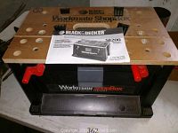 Front view of the Black & Decker Workmate ShopBox Portable Project Center showing wooden top with holes, red clamps, black plastic base, and instruction manual placed on top.