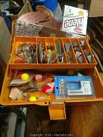 Photo showing full view of an orange fishing tackle box open with multiple compartments holding fishing hooks, metal fishing lures, sinkers, bobbers, packaging for a fishing scale, and fishing line.