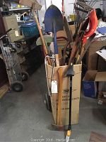Photo showing a variety of lawn and garden tools including shovels, rakes, hoes, and pruning saw in a cardboard box next to a dolly, tools have different handle materials and colors.