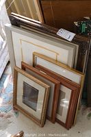 Photo showing a grouping of various picture frames stacked leaning against wall on carpeted floor.