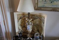 Full view of lamp with ceramic cherubs and floral decorations sitting in front of a vintage map wallpaper, surrounded by various beer steins and mugs (not included).
