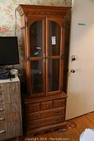 Full view of the tall wooden cabinet with glass doors and drawers beside a dresser and a door.