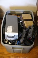 Top view of plastic storage bin filled with various cameras, camcorder, and a film scanner. Accessories and cases visible.