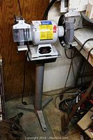 Front view of the bench grinder mounted on the metal stand, showing both grinding wheels, protective shields, and tool rests.