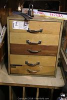 Full view of the wooden cabinet with four drawers, metal handles, and a hasp lock secured on top without a key. Visible wear on the cabinet surface.