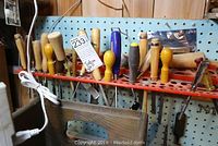 Photo showing a set of vintage chisels with wooden handles held in a red rack against a pegboard wall with a coil of white electrical cord onsite.