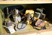 Dial indicator micrometer and several power tools including a yellow belt sander, a Bosch palm sander, and a green hand-held tool on a shelf with tags.