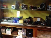Overview of shelves displaying vintage cameras including Keystone, Konica, and Ansco; boxed accessories below
